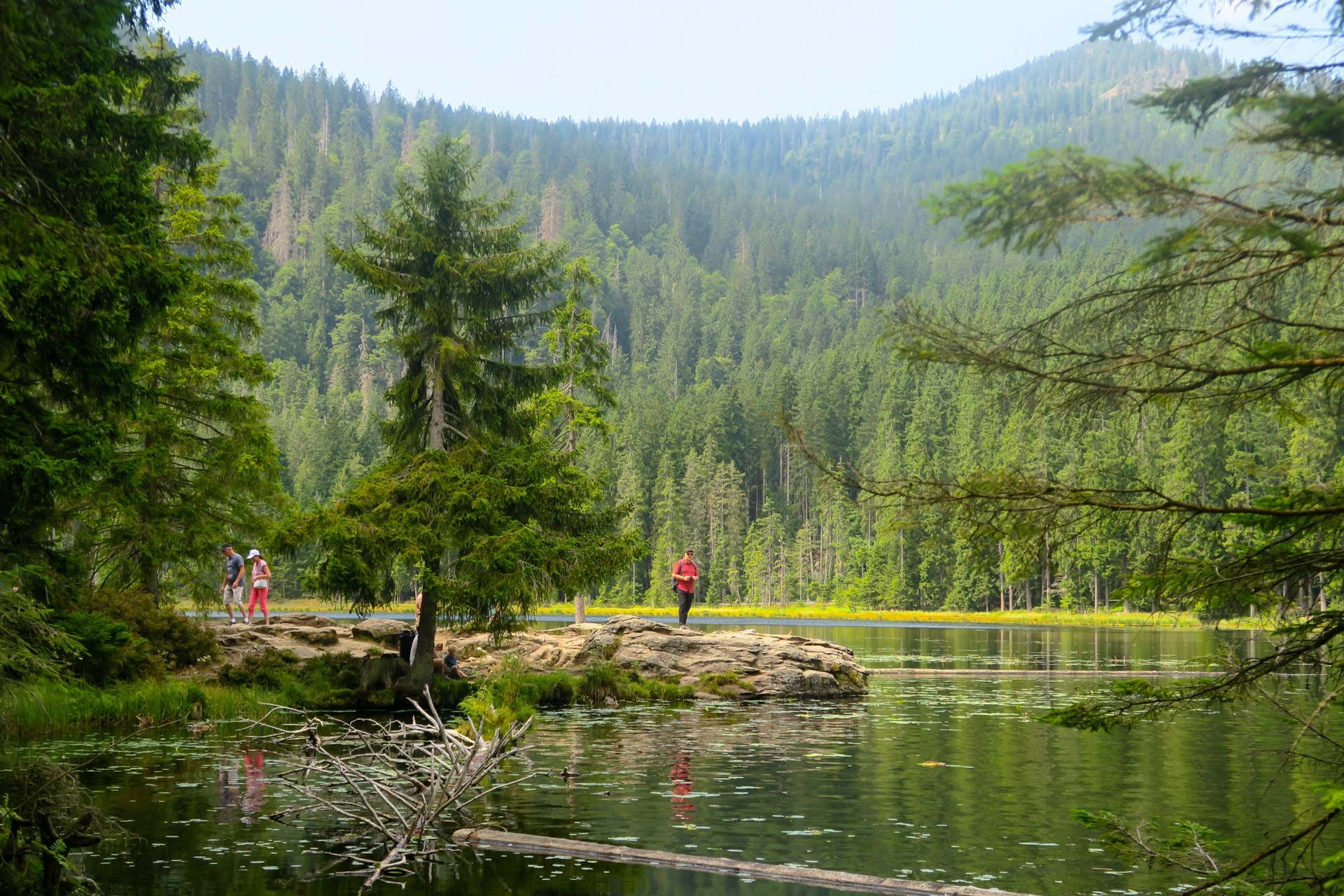eine Felseninsel im See, zwei Wanderer links und ein rot gekleideter Fotograf direkt auf dem Felsen